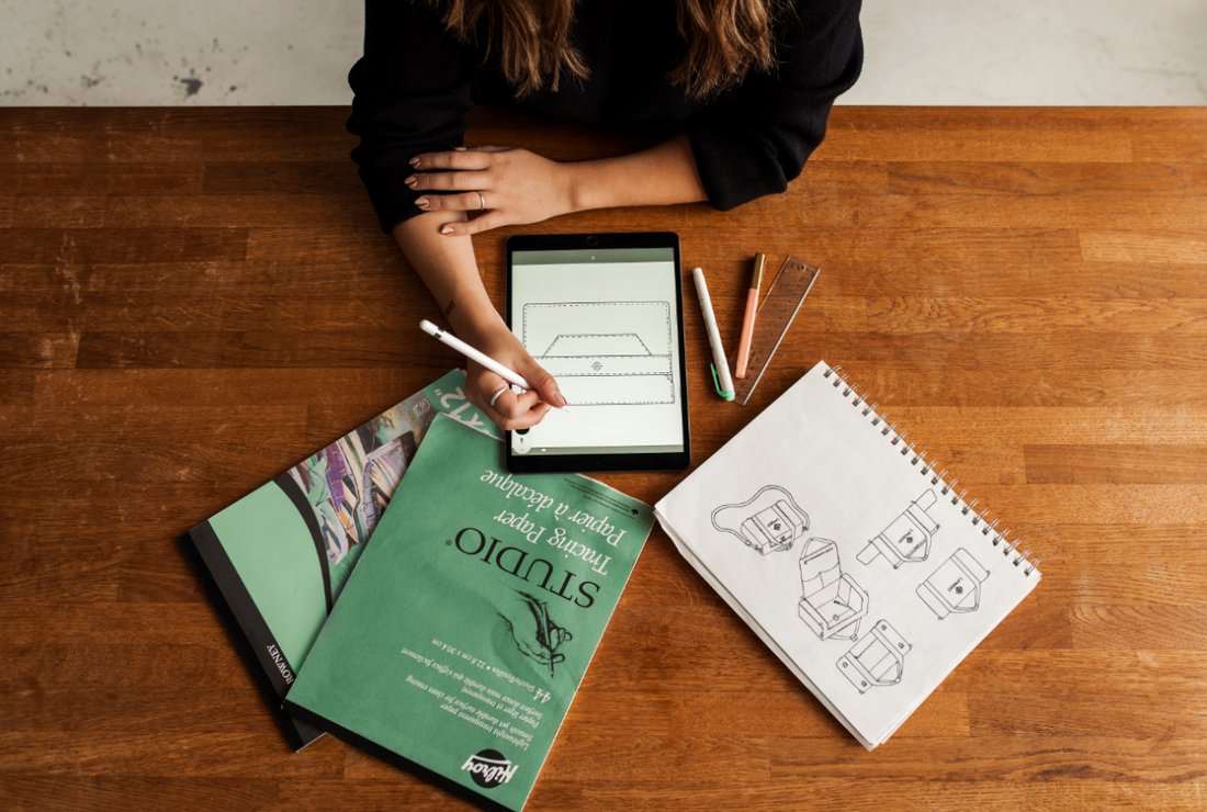 Overhead view of a designer sketching a handbag on a tablet, with bag drawings, art paper, and drawing tools on a wooden table.
